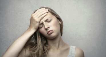 A woman with long hair holds her forehead, eyes closed, appearing to have a headache. She wears a gray tank top against a plain background.