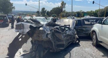 A severe car crash at a busy intersection with a heavily damaged black car at the center. Multiple vehicles are involved, with emergency personnel on site.
