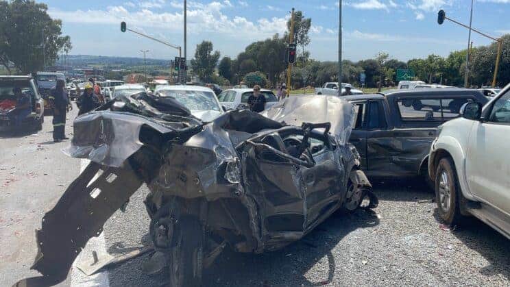 A severe car crash at a busy intersection with a heavily damaged black car at the center. Multiple vehicles are involved, with emergency personnel on site.