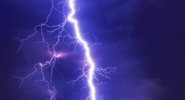 Bright lightning bolt splitting a dark purple night sky, illuminating surrounding clouds. The scene conveys a dramatic and intense atmosphere.