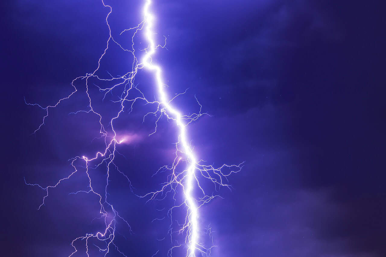 Bright lightning bolt splitting a dark purple night sky, illuminating surrounding clouds. The scene conveys a dramatic and intense atmosphere.