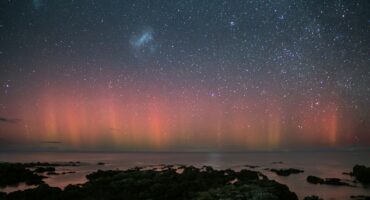 Starry night sky above a rocky coastline with shimmering aurora in shades of red and orange. Reflective water and serene ambiance capture the ethereal beauty.