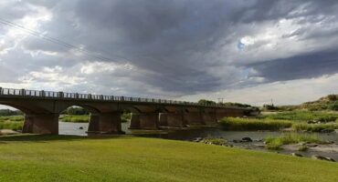 A long concrete bridge crosses over a calm river with green grassy banks. Dark, cloudy sky hints at a storm, adding a dramatic and moody atmosphere.