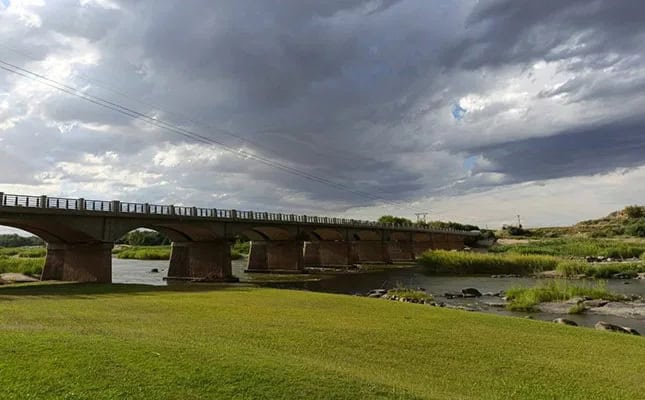 A long concrete bridge crosses over a calm river with green grassy banks. Dark, cloudy sky hints at a storm, adding a dramatic and moody atmosphere.