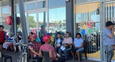 A group of people sit patiently outside a building with large windows, some holding papers. The mood is calm. A colorful handprint design is on the glass.