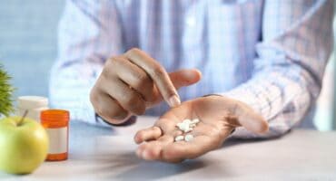 A person in a plaid shirt holds several white pills in their palm, pointing with the other hand. Nearby are an apple, a plant, and an open pill bottle, suggesting a focus on health.