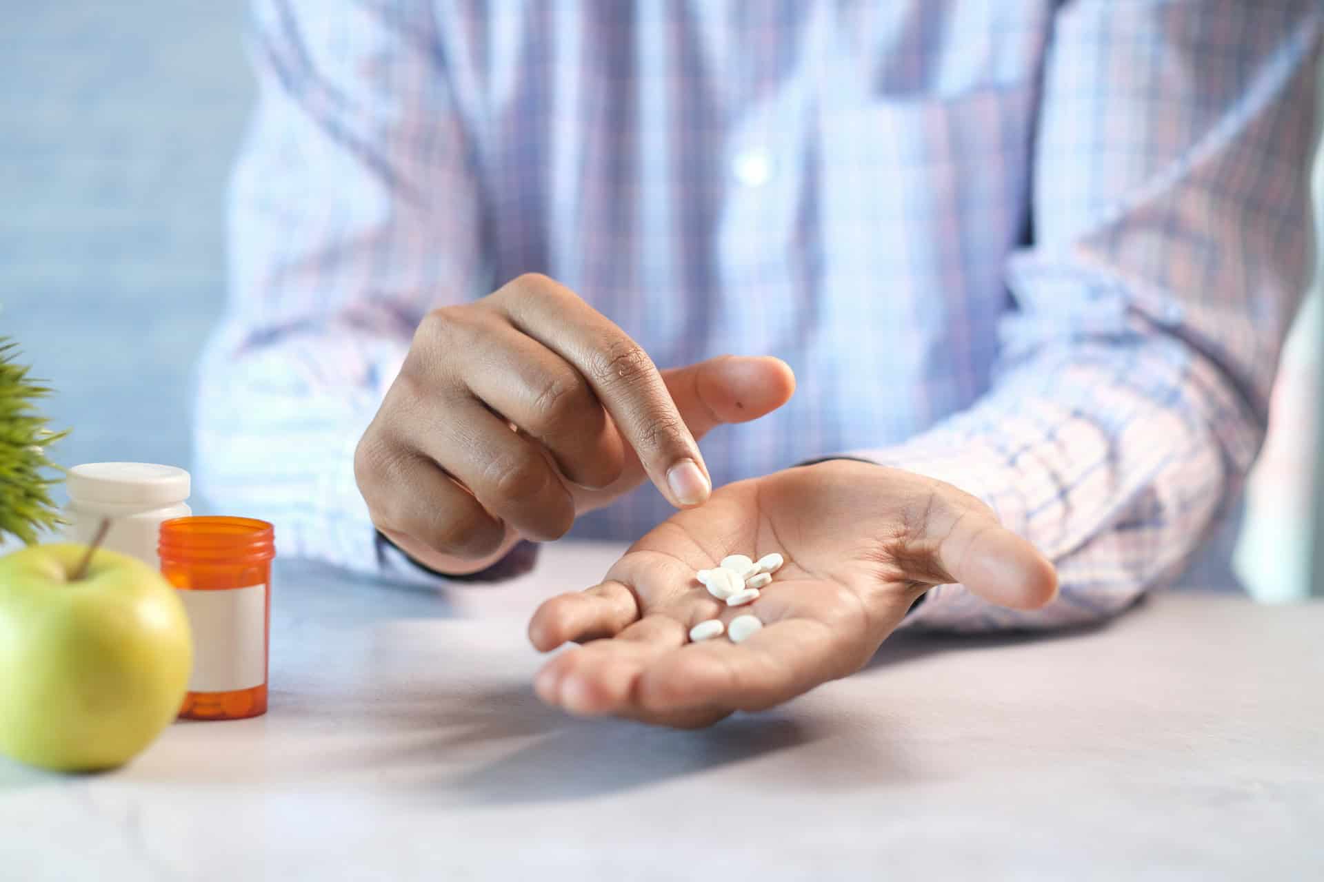 A person in a plaid shirt holds several white pills in their palm, pointing with the other hand. Nearby are an apple, a plant, and an open pill bottle, suggesting a focus on health.