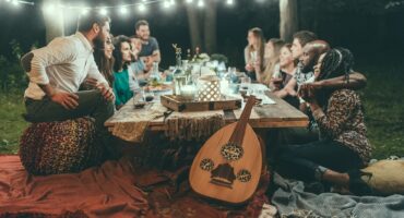 A group of people sit around a low wooden table outdoors at night, under string lights, on colorful cushions and blankets. A lantern and an oud are visible, creating a cozy, festive atmosphere.