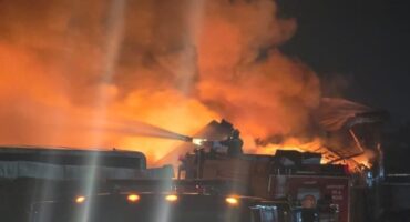 Flames and smoke turn a dark sky orange with fire trucks visible in front of the blaze