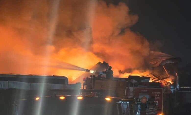 Flames and smoke turn a dark sky orange with fire trucks visible in front of the blaze
