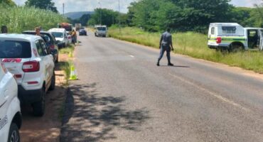 Crime scene next to a tar road with cones visible on the left and a police officer crossing the road to a police vehicle standing on the right-hand side of the road.