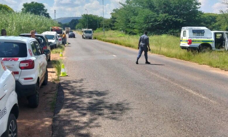 Crime scene next to a tar road with cones visible on the left and a police officer crossing the road to a police vehicle standing on the right-hand side of the road.