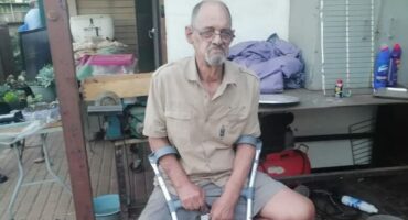 Man with crutches dressed in a khaki shirt sitting on a chair.