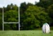 White Rugby ball visible on a green field with goal posts in the background.