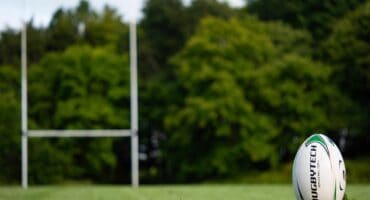 White Rugby ball visible on a green field with goal posts in the background.