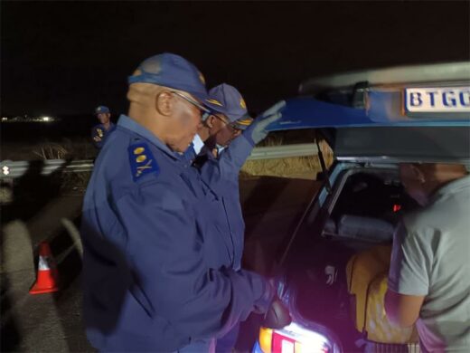 Three uniformed officers inspect a vehicle's trunk at night on a roadside, under dim lighting, conveying a serious, focused atmosphere.