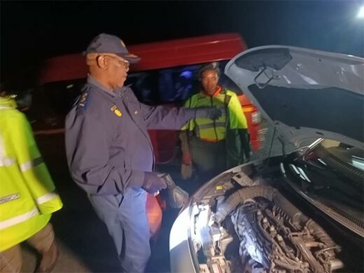 A police officer inspects a car engine at night, using a flashlight. Nearby, a police van is illuminated and another officer observes, creating a tense yet focused scene.
