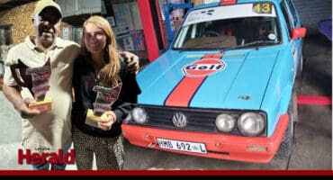 Peter Pilusa and Clarisse Lourens proudly hold the trophies in front of a blue and red Volkswagen Golf racing car marked with the number 13 and a “Golf” logo.