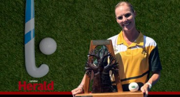 Ashley van Schalkwyk holds a trophy. A hockey stick and ball are visible in the background.