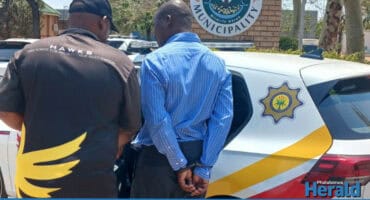 A Hawks officer arrests senior offical outside the Maruleng Local Municipality building, guiding him toward a marked police vehicle parked nearby.