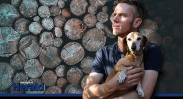 Deon Breytenbach from Bushveld Turnery holds his dachshund while standing in front of a background featuring stacked logs, representing his woodturning craft.