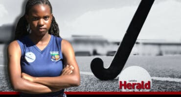 Buhle stands cross-armed and serious. In the background, a hockey stick and ball in black and white.