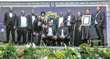 A group of men and women dressed in formal black attire pose on stage during an award ceremony. One woman holds a framed certificate while another person displays a trophy. The background features the South African Police Service emblem and stage lighting, with plants arranged in front of the stage.