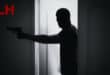 A masked man holding a gun walks through a door.