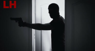 A masked man holding a gun walks through a door.