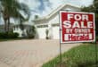 a sign in front of a house that has 'for sale' written on it, the house at the back is blurred slightly with a brick driveway leading toward the garages, some trees and foliage are next to the house and the house itself has an A-frame roof