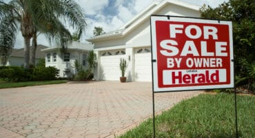 a sign in front of a house that has 'for sale' written on it, the house at the back is blurred slightly with a brick driveway leading toward the garages, some trees and foliage are next to the house and the house itself has an A-frame roof