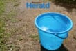 a blue bucket filled with clean water on a gravel path with some green grass.
