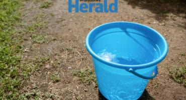 a blue bucket filled with clean water on a gravel path with some green grass.