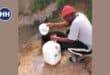 a person wearing a white shirt, black pants and an orange hat bends over with buckets in hand to collect water from a well in the ground
