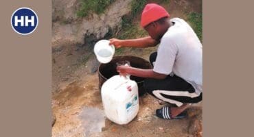 a person wearing a white shirt, black pants and an orange hat bends over with buckets in hand to collect water from a well in the ground