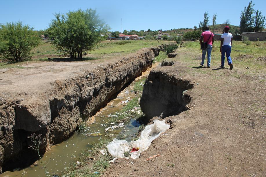 Residents walk past the ditch that has washed out as a result of the water and sewage streaming through Wedela