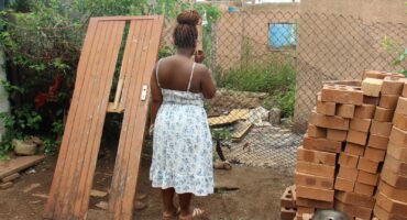 Nandipha Rudaza looks into her neighbouring yard, where criminals are regularly hiding in the house damaged by the sinkhole. She is standing next to the door of her family's house that was smashed by the criminals when they broke in.