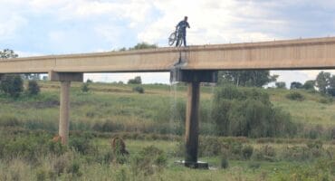 A cyclist carrying his bicycle over the bridge from where sewage is cascading from a municipal pipeline onto the ground next to the river.