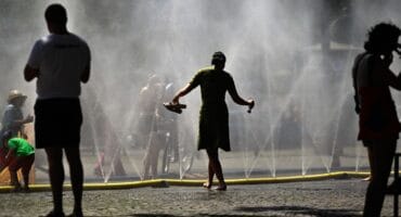 People stand near over water hoses spilling water to provide relief from the summer heat as part of Cologne’s climate adaptation measures in Cologne, western Germany on June 25, 2026 during a heatwave in Europe.