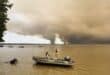 A beachgoer walks in Lake Lacanau, also called Etang de Lacanau (Lacanau Pond) from the beach of Moutchic in Lacanau, southwestern France on July 24, 2026, as clouds of smoke rise into the sky from a wildfire. Photo: AFP