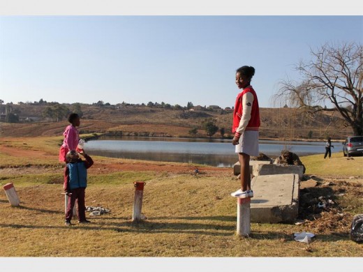On the right, Ashley Mongwe climbs a post while playing next to Coronation Dam. Mesho Mongwe is drinking some cooldrink and Tiyani joins Ashely on the post.
