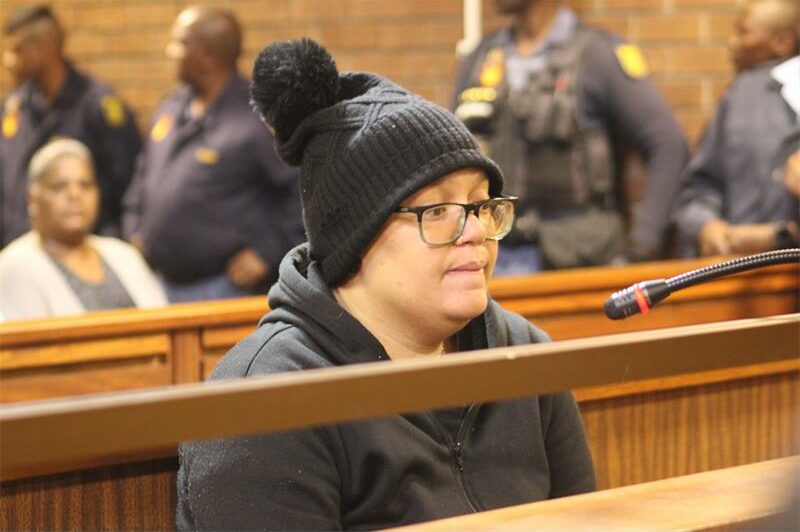 A person wearing glasses and a black beanie sits in a courtroom, appearing serious. Police officers stand in the background, creating a tense atmosphere.