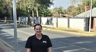 A woman stands on a suburban street next to a triangular road sign indicating a speed bump. She is smiling, wearing a black shirt and pants. The scene is calm and sunny.