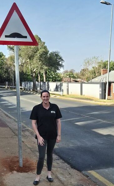 A woman stands on a suburban street next to a triangular road sign indicating a speed bump. She is smiling, wearing a black shirt and pants. The scene is calm and sunny.