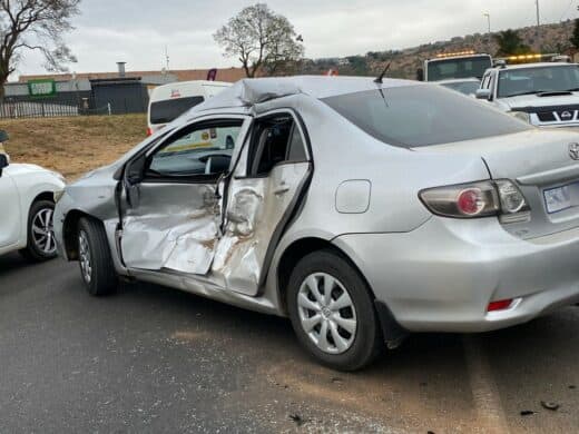 A silver sedan with heavy damage to the driver's side is parked on the road after a collision, surrounded by other vehicles and tow trucks.