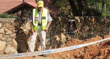 Man wearing a yellow cap and neon safety vest inspects a construction excavation site in front of a stone wall. The ground is dug up with soil piles marked off by white barrier tape labeled “Joburg Water.” Behind him, a warning sign on the wall reads “Warning Armed Response” with an image of a guard dog. The background shows a house with a tiled roof, trees, and garden plants.