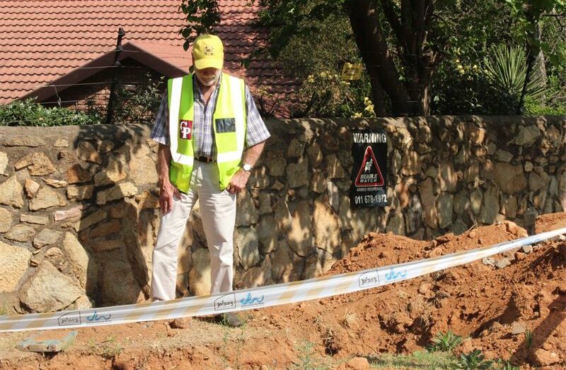 Man wearing a yellow cap and neon safety vest inspects a construction excavation site in front of a stone wall. The ground is dug up with soil piles marked off by white barrier tape labeled “Joburg Water.” Behind him, a warning sign on the wall reads “Warning Armed Response” with an image of a guard dog. The background shows a house with a tiled roof, trees, and garden plants.
