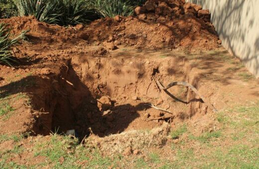 Large hole dug into the ground with exposed soil and broken underground pipes visible inside the excavation site. Piles of reddish-brown dirt are heaped at the back, while patches of green grass remain around the edges. The hole is positioned near a stone or concrete wall, with plants and shrubs in the background.