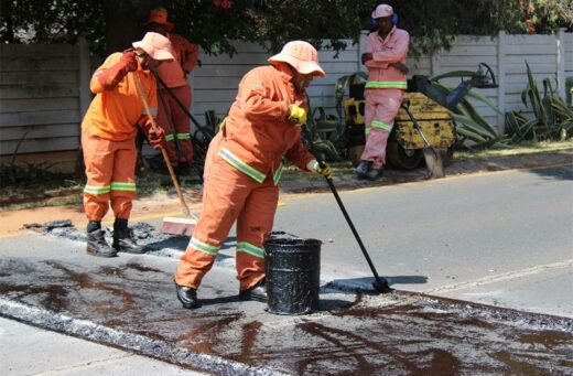 Road workers in orange uniforms and hats apply tar to a road, smoothing it with tools. A compactor is in the background.