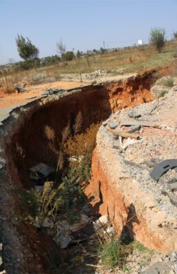A large, eroded pit in dry ground with exposed orange soil and scattered debris. Sparse vegetation and trees are visible under a clear blue sky.